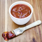 Small bowl of sweet and spicy BBQ sauce with a brush full of sauce in front of it.