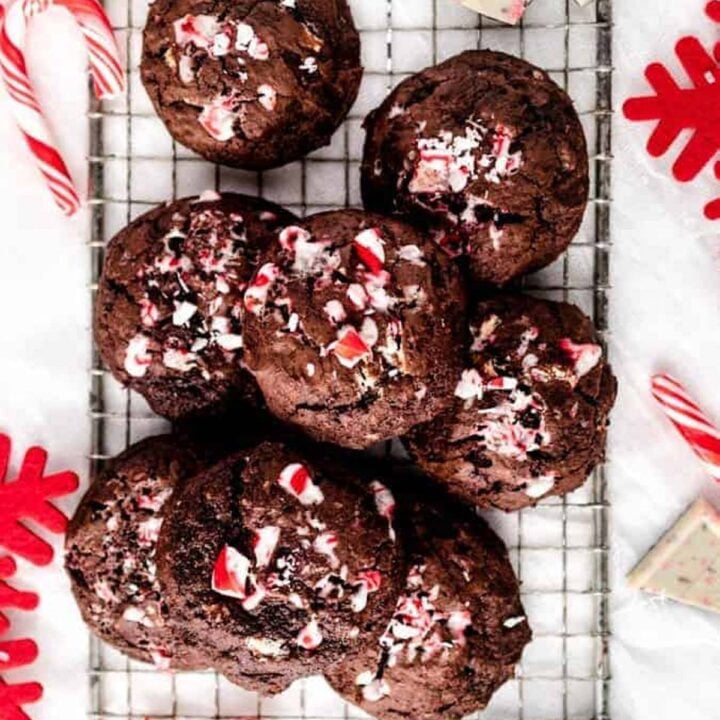 Baked chocolate cookies topped with crushed candy cane sit on a wire cooling rack, surrounded by mini candy canes, squares of white chocolate, and red snowflake decorations.