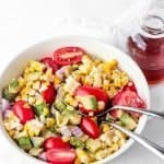 Avocado Corn Tomato salad with red onion ready to be eaten with two spoons in the bowl and dressing on the side ready to be poured in