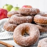 pile of baked apple cider donuts on a wire rack with apples in the background and cinnamon sticks in front