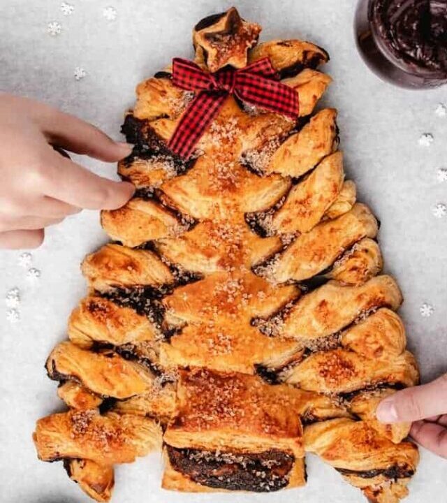 Overhead of puff pastry Christmas tree with red and black bow and two hands grabbing for the pastry