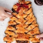 Overhead of puff pastry Christmas tree with red and black bow and two hands grabbing for the pastry