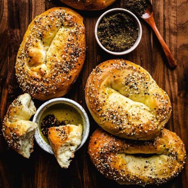 Five oval bagels covered in everything seasoning sit on a dark wood board next to a bowl of za'atar and a dish of olive oil with torn pieces of bagel.