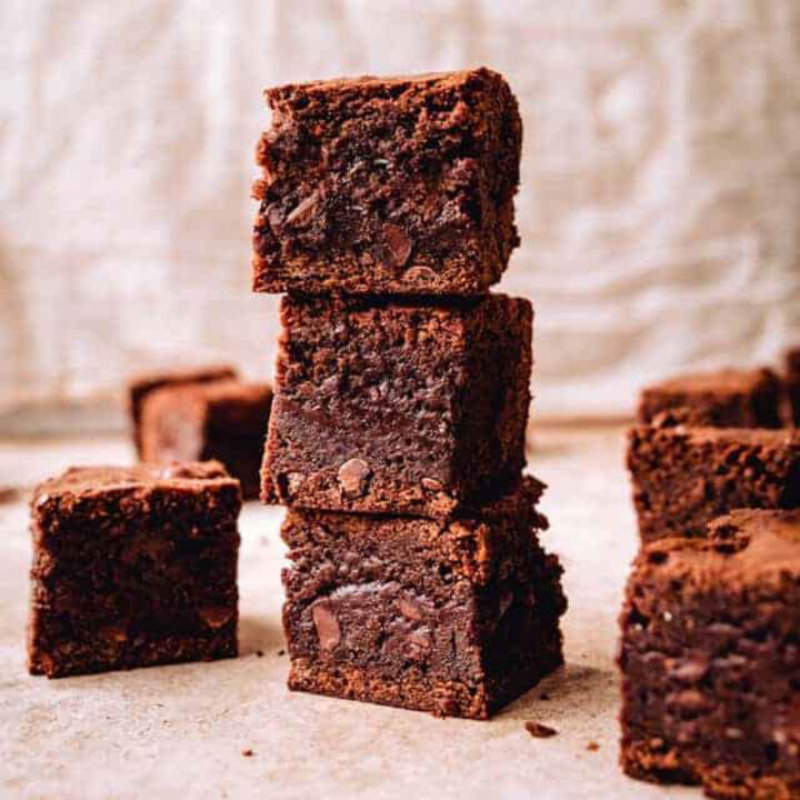 A tall stack of three thick, chewy chocolate brownies with more brownie squares around it on a parchment-colored surface.