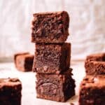 A tall stack of three thick, chewy chocolate brownies with more brownie squares around it on a parchment-colored surface.