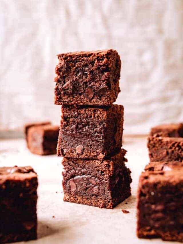 A tall stack of three thick, chewy chocolate brownies with more brownie squares around it on a parchment-colored surface.