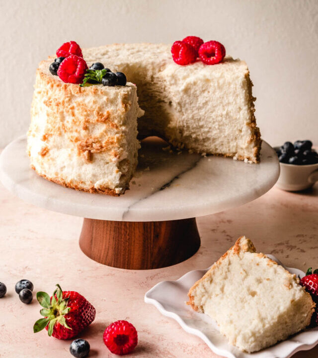 Angel food cake on a cake stand with berries on top and a slice removed, served on a plate in front.