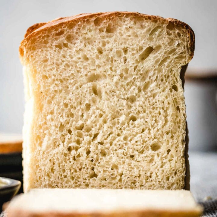A tall loaf of white bread cut open to show an airy crumb full of holes, with a thick slice lying in front of it.