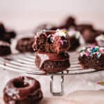 Mini chocolate donuts stacked with a bite missing out of one and other mini donuts on the cooling rack around it.