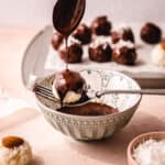 A fork holding a coconut ball over a bowl of chocolate and a spoon drizzling chocolate over it.