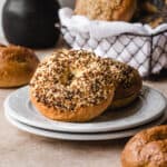 Two bagels laying on a plate with a basket of bagels behind it.