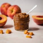 Small pieces of peach around a glass jar of peach jam.