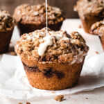 Glaze being drizzled over a cinnamon streusel muffin on parchment paper.