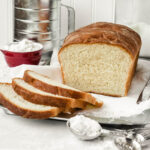 Tablespoon of flour sitting in front of a loaf of Amish white bread with a few slices laying next to it.