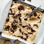 Ice cream scoop resting in a square baking dish filled with s'mores ice cream topped with chocolate chunks.